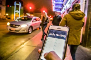 Imagen de la mano de una persona caminando por la calle, sosteniendo un smarthphone que se ve en la pantalla una app de ride hailing - Diferencias entre el car-sharing y el ride-hailing